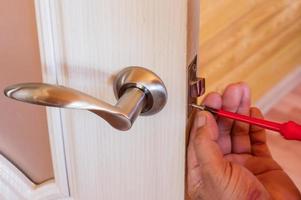 faz-tudo repara a fechadura da porta na sala, homem fixando a fechadura com chave de fenda, close-up da porta de reparação, instalação de serralheiro profissional foto