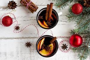 vinho quente em canecas esmaltadas brancas com especiarias e frutas cítricas na mesa de madeira com galhos de árvores de peles e presentes de natal. foto