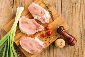 bife cru de carne de porco jovem em uma placa de cozinha e uma mesa de madeira com cebola verde, alecrim, tomate. foto
