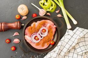 bife cru, entrecosto com especiarias para cozinhar carne em uma panela de ferro fundido com cebola, alho e tomate. fundo preto. vista do topo. foto