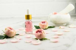 frasco de vidro com pipeta rosa cosmético para cuidados com a pele facial antienvelhecimento em uma mesa de mármore com botões de rosa e pétalas. foto