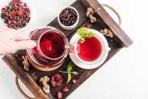 cerimônia do chá. a mão de uma mulher derrama chá de hibisco vermelho perfumado em uma xícara de folhas de hortelã. vista superior da bandeja de madeira com chá da manhã. foto