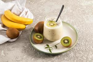 smoothie de frutas energéticas feito de bananas e kiwi em um copo em uma placa de cerâmica e um fundo de concreto com frutas. o conceito de alimentação saudável. foto