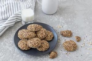 biscoitos de aveia com sementes de linho, gergelim e passas em um prato escuro e um copo de leite em um fundo cinza. foto