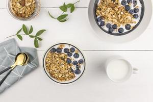 vista superior de três tigelas de granola assada e leite fresco, mirtilos e nozes. café da manhã delicioso e saudável. fundo de madeira branco. foto