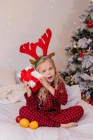 menina loira feliz em casa de pijama para o natal classificando presentes e brincando com brinquedos de natal foto