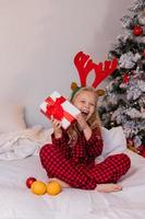 menina loira feliz em casa de pijama para o natal classificando presentes e brincando com brinquedos de natal foto