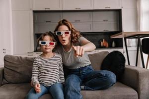 garota de jeans e sua filha estão assistindo filme em 3d, sentado em casa no sofá foto