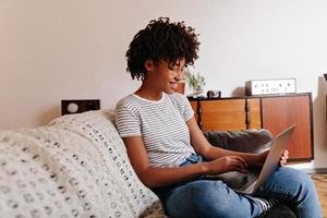 garota africana em camiseta listrada está sentada no sofá e conversando com um sorriso no laptop. foto
