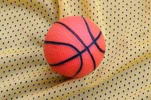 pequena bola de basquete de borracha laranja encontra-se em uma textura de tecido de roupa de camisa esportiva amarela e fundo com muitas dobras foto