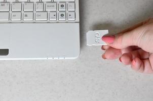 uma mão feminina insere um cartão SD compacto branco na entrada correspondente na lateral do netbook branco. mulher usa tecnologias modernas para armazenar memória e dados digitais foto