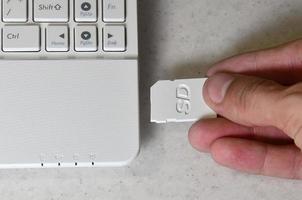 uma mão masculina insere um cartão SD compacto branco na entrada correspondente na lateral do netbook branco. homem usa tecnologias modernas para armazenar memória e dados digitais foto