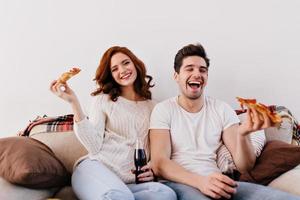 amigos bem-humorados posando no sofá aconchegante. homem de camiseta branca comendo pizza com a namorada.