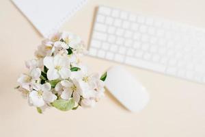 teclado, folha de papel branca e mouse e flores de maçã branca em um vaso fora de foco em uma mesa bege. conceito de layout e design. postura plana. foto
