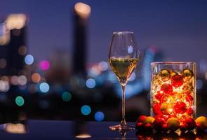 único copo de vinho branco que tem enfeites de natal com luzes em vaso de vidro sobre fundo claro bokeh cidade colorida. foto