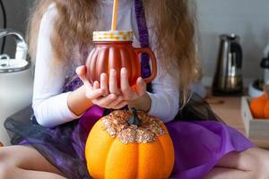 criança bebe suco de abóbora, smoothie de vegetais ou coquetel de uma caneca com um canudo. cozinha decora para o halloween. garota em uma fantasia de bruxa. outono aconchegante em casa