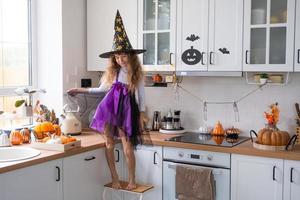 criança decora a cozinha em casa para o halloween. garota em uma fantasia de bruxa brinca com a decoração para o feriado - morcegos, lanterna jack, abóboras. conforto de outono em casa, cozinha em estilo escandinavo, loft foto