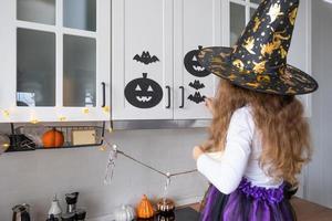 criança decora a cozinha em casa para o halloween. garota em uma fantasia de bruxa brinca com a decoração para o feriado - morcegos, lanterna jack, abóboras. conforto de outono em casa, cozinha em estilo escandinavo, loft foto