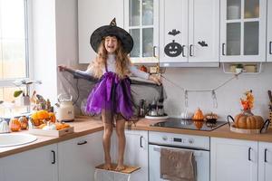 criança decora a cozinha em casa para o halloween. garota em uma fantasia de bruxa brinca com a decoração para o feriado - morcegos, lanterna jack, abóboras. conforto de outono em casa, cozinha em estilo escandinavo, loft foto