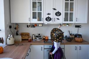 criança decora a cozinha em casa para o halloween. garota em uma fantasia de bruxa brinca com a decoração para o feriado - morcegos, lanterna jack, abóboras. conforto de outono em casa, cozinha em estilo escandinavo, loft foto