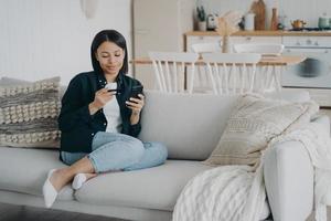 serviço bancário on-line. mulher segurando cartão de crédito, telefone faz pagamento sem dinheiro no sofá em casa foto