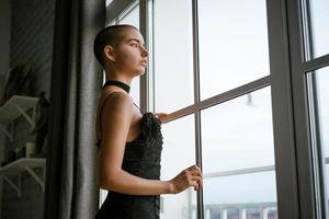 retrato de uma jovem triste com cabelo curto em pé olhando pela janela em um vestido preto foto