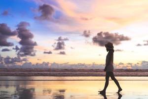 vista traseira de uma jovem turista asiática caminhando relaxando na areia da praia com um belo céu dramático do pôr do sol foto