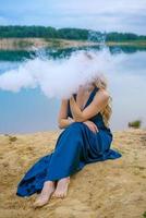 jovem de vestido azul senta-se na margem de um lago durante o dia. uma nuvem de fumaça antes foto