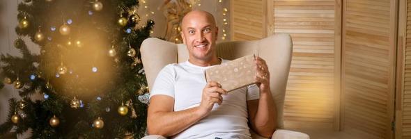 homem feliz na cadeira no fundo da árvore de natal com um presente nas mãos foto