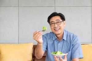 homens asiáticos seniores sentados no sofá em casa enquanto comem salada de vegetais verdes e suco de laranja orgânico no café da manhã. estilo de vida saudável para o conceito de pessoas de aposentadoria mais velha. foto