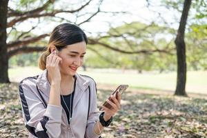 foco seletivo no rosto de jovens mulheres asiáticas bonitas usando relógio inteligente para rastrear a atividade e ouvir música do smartphone enquanto se aquece antes do exercício de ioga com natural e árvores ao fundo. foto