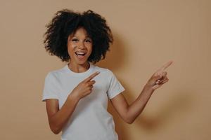 jovem garota afro-americana sorrindo alegremente e apontando com o dedo indicador para o lado foto