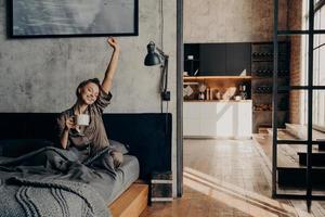 jovem mulher bonita sentada na cama esticando com a mão para cima enquanto segura a xícara de café foto