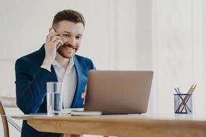 jovem executivo elegante falando no telefone na frente do laptop atrás da mesa de escritório foto