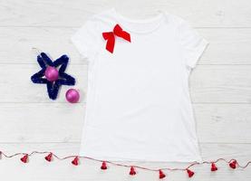 feche a camiseta branca modelo em branco com espaço de cópia e conceito de férias de natal. t-shirt de maquete de vista superior e laço vermelho sobre fundo branco de madeira. feliz ano novo acessórios de decorações. roupa de natal foto