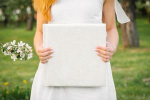 um álbum de fotos de casamento coberto de branco é segurado por uma mulher de vestido branco