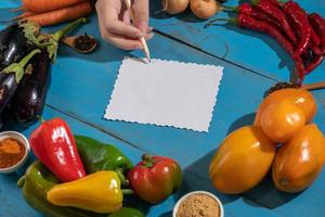 legumes são dispostos em torno de uma folha de papel e um lápis. espaço vazio para texto. legumes, vazio em branco para receita em um fundo azul. foto