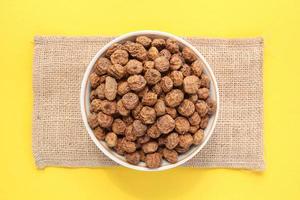 Chufa, Tigernut, amêndoa da terra na tigela de cerâmica branca e saco de lona eco perto de colher de pau em fundo amarelo. conceito de comida vegana saudável. noz de tigre para farinha, óleo, leite, bebida tradicional horchata foto