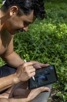 yogi masculino bonito ativo desenho tablet andando na floresta antes ou depois de um treino. aula de ioga jovem hispânico atlético com um tapete de fitness foto