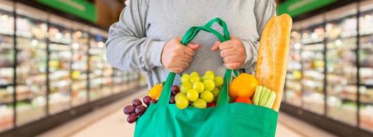cliente segura sacola de compras verde reutilizável com frutas e vegetais com fundo de corredor de supermercado foto