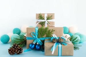 três caixas de presente de natal embrulhadas em papel artesanal, fitas azuis e brancas, decoradas com ramos de abeto, pinhas e bolas de natal no fundo azul e branco com luzes de natal. foto