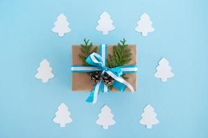 caixa de presente embrulhada de papel artesanal, fitas azuis e brancas e ramos de abeto decorados e pinhas no fundo azul com árvore de abeto de papel branco, vista superior. presente de Natal. foto