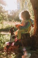 menina em uma fantasia de halloween de unicórnio arco-íris e um dachshund em um vestido com uma cesta de abóbora para doces estão sentados no toco ao pôr do sol da floresta. fabulosa maravilhosa floresta mágica. espaço para texto foto