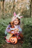 menina em uma fantasia de halloween de unicórnio arco-íris e um dachshund em um vestido com uma cesta de abóbora para doces estão sentados no toco ao pôr do sol da floresta. fabulosa maravilhosa floresta mágica. espaço para texto foto