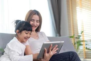 linda mãe asiática com filha sentada no sofá e jogando videogame em um tablet enquanto passam tempo juntos em casa
