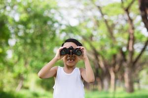 garotinho de pé em um campo gramado verde, examinando a floresta circundante com binóculos enquanto explora o campo foto