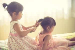linda garotinha escovando o cabelo de sua irmã enquanto está sentado na cama foto