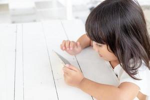 menina asiática jogando smartphone em cima da mesa. assistindo smartphone. as crianças usam telefones e jogam. criança usa celular. jogo viciante e desenho animado foto