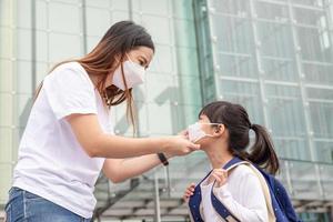 mãe asiática ajuda a filha a usar uma máscara médica para proteção covid-19 ou surto de coronavírus a se preparar para ir à escola quando voltar à ordem escolar. foto