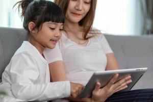 linda mãe asiática com filha sentada no sofá e jogando videogame em um tablet enquanto passam tempo juntos em casa